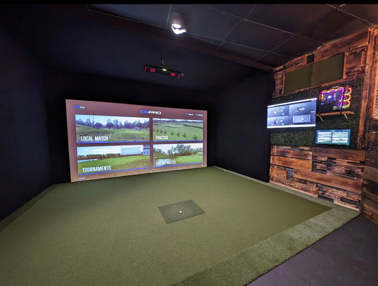 Golf simulator built inside a barn, with a projected course on the impact screen, a turf hitting area, wood-panel accent wall, and wall-mounted displays