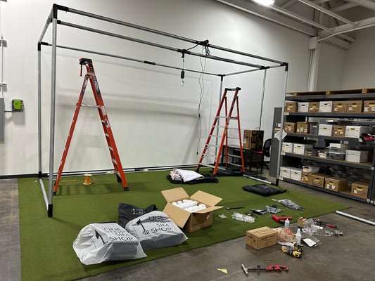 Steel-tube golf simulator enclosure frame being assembled over green turf, with ladders and boxed parts on the floor