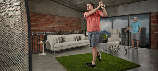 Golfer swinging off a turf hitting mat in a home living space with a Garmin Approach R10 launch monitor behind the ball