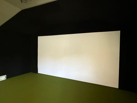 Taut white golf impact screen mounted in a black simulator enclosure above green turf flooring