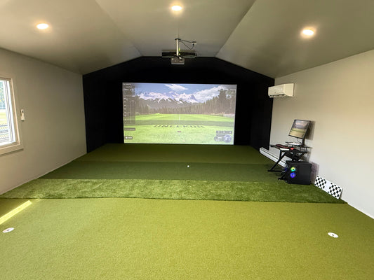 Finished home golf simulator room with a course projected on the impact screen, ceiling projector, turf floor, and a PC station along the wall